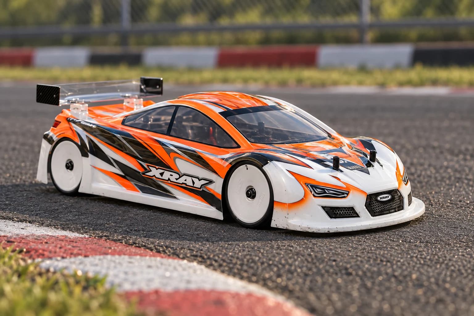 An orange, white and graphite RC touring car beside the kerb of an asphalt circuit